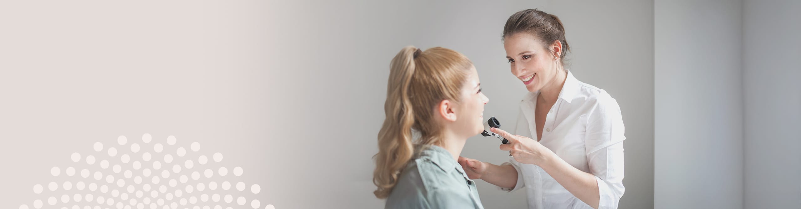A female dermatologist smiling while performing a skin examination on a young patient using a dermatoscope.